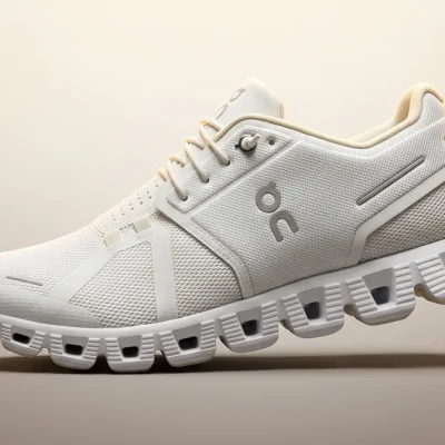 Premium photoreal product close-up of a single white-and-cream On Cloud running shoe with its distinctive CloudTec sole pods, three-quarter side view on a clean warm cream-to-taupe studio gradient backdrop, with a small ON brand logo lockup in the upper-left corner