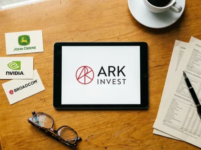 Overhead view of a wooden desk with a tablet showing the ARK Invest logo, printed cards bearing the John Deere, Nvidia and Broadcom logos, a pen on report pages and a coffee cup