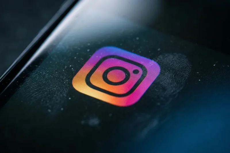 Extreme close-up of the Instagram icon glowing on a dark smartphone screen, with a fingerprint smudge and dust visible on the glass beside it
