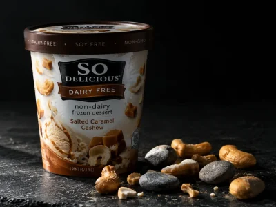 Macro photograph of a So Delicious Dairy Free non-dairy frozen dessert pint on dark slate, with caramel cashew pieces and several small grey stones scattered beside it