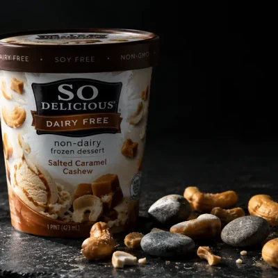 Macro photograph of a So Delicious Dairy Free non-dairy frozen dessert pint on dark slate, with caramel cashew pieces and several small grey stones scattered beside it