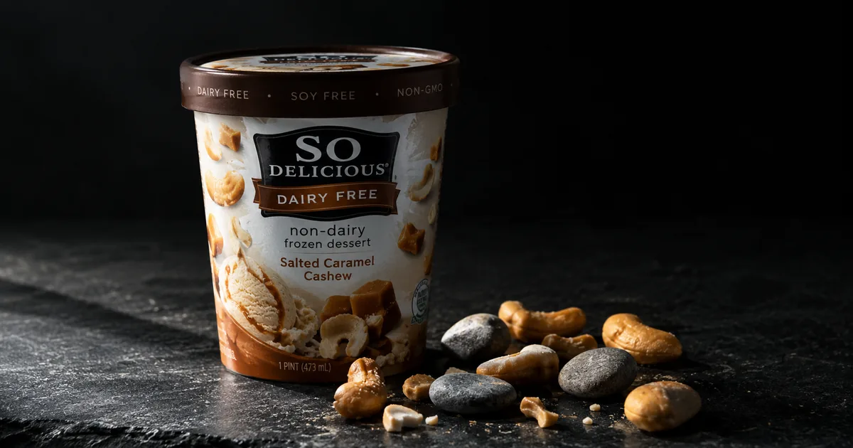 Macro photograph of a So Delicious Dairy Free non-dairy frozen dessert pint on dark slate, with caramel cashew pieces and several small grey stones scattered beside it