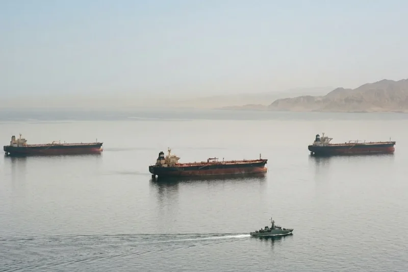 Hazy wide shot of three laden crude oil supertankers sitting at anchor in the Strait of Hormuz while a small naval escort vessel passes in the foreground