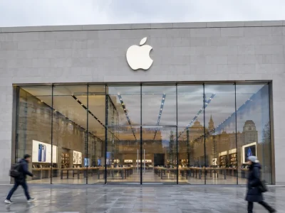Exterior of an Apple Store on an overcast day, the Apple logo mounted on the stone facade above a full-height glass frontage as pedestrians walk past