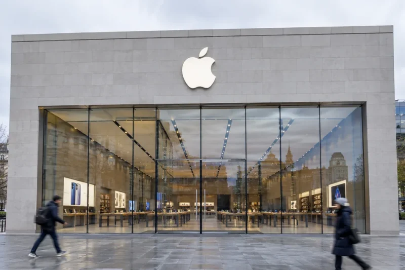 Exterior of an Apple Store on an overcast day, the Apple logo mounted on the stone facade above a full-height glass frontage as pedestrians walk past