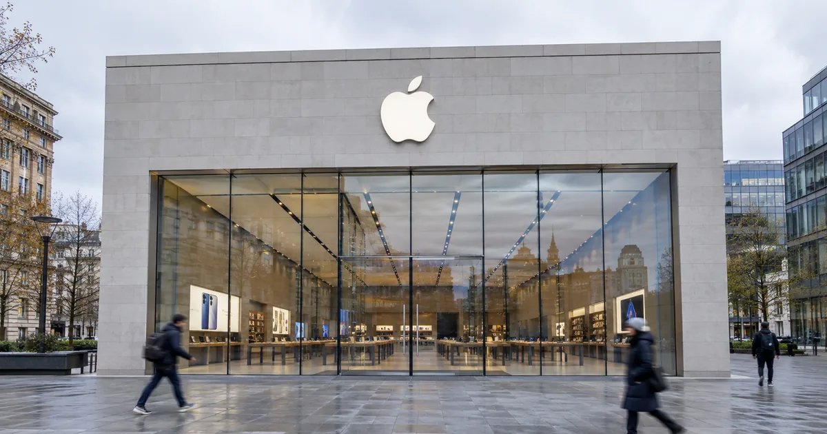Exterior of an Apple Store on an overcast day, the Apple logo mounted on the stone facade above a full-height glass frontage as pedestrians walk past