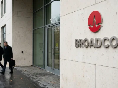 Red Broadcom logo and wordmark on a pale stone wall beside a glass office entrance as two workers walk past on wet pavement