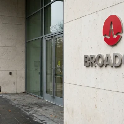 Red Broadcom logo and wordmark on a pale stone wall beside a glass office entrance as two workers walk past on wet pavement