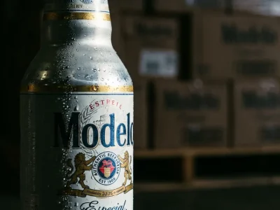 Close-up of a cold Modelo Especial bottle with condensation, stacked beer cases on a warehouse pallet blurred behind it