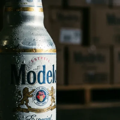 Close-up of a cold Modelo Especial bottle with condensation, stacked beer cases on a warehouse pallet blurred behind it