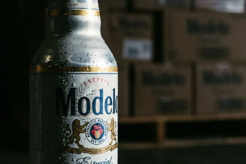 Close-up of a cold Modelo Especial bottle with condensation, stacked beer cases on a warehouse pallet blurred behind it