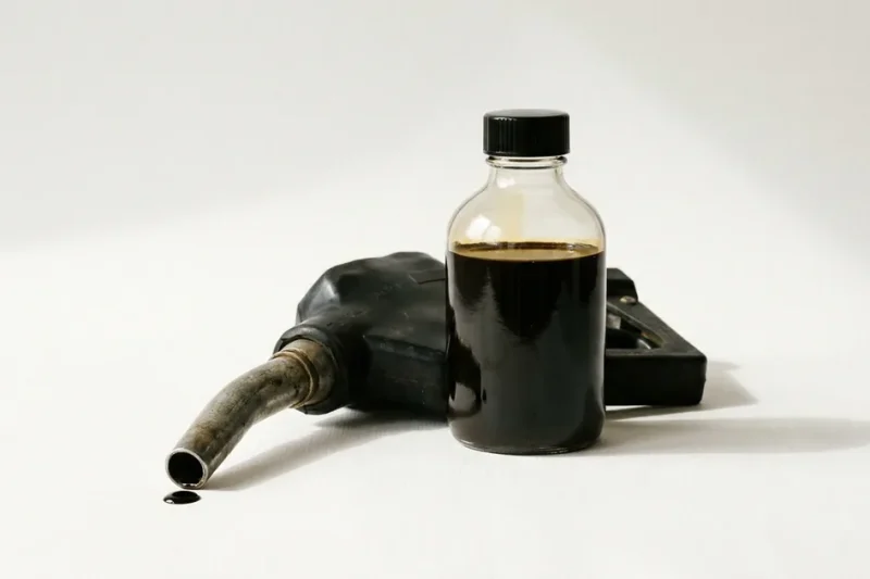 A glass sample bottle of dark crude oil next to a worn gasoline pump nozzle on a white studio backdrop, with a drop of oil on the paper
